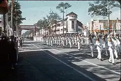 Un homme asiatique et un groupe d'hommes blancs, se tenant debout sur un terrain d'aviation militaire, dans une position de recueillement.