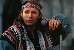 Photographie en couleur, mi-buste d'un homme, regard dirigé sur la gauche, avec bandeau sur le front, cheveux longs, bras gauche appuyé sur un micro sur pied, vêtu d'un poncho à motifs géométriques colorés.