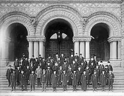 Photo en noir et blanc d'un groupe d'homme se tenant debout dans un escalier devant la façade d'un bâtiment composée de trois arches
