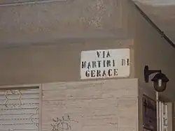 Photographie d'un angle de rue en plan rapproché avec peint sur le mur un rectangle blanc, avec inscrit dessus en lettres majuscules noires : Via Martiri di Gerace.