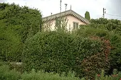 Photo en couleur d'un bout de façade claire sous un toit pointu émergeant de massifs d'arbres et de verdure.