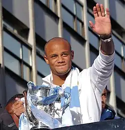 Vincent Kompany, de face, lors de la célébration de fin de saison 2013-2014. Il tient le trophée de la Coupe de la ligue d'Angleterre dans la main droite et fait un signe à la foule de la main gauche.