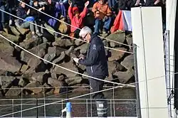 Cheveux blancs, lunettes, de profil. Debout sur son bateau, salué par la foule dans le chenal des Sables-d'Olonne, il prépare en souriant son téléphone portable pour filmer le public.