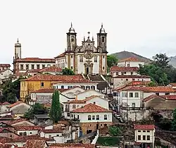 Photographie en couleur. Vue panoramique de la vieille ville d'Ouro Preto avec des maisons blanches au toit de tuiles rouges surplombées d'une église blanche en haut d'une colline.