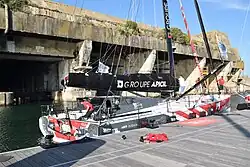 Vu de trois quarts arrière, voilier rouge, blanc et gris amarré au ponton, devant la base de sous-marins de Lorient.