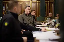 Portrait d'un homme blanc, assis à une table avec d'autres hommes blancs. La table est parsemée de documents.