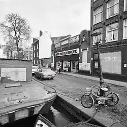Cliché en noir et blanc d'un alignement de bâtiments, où domine la brique, le long d'un canal : l'un d'eux est une ancienne salle de cinéma de style mauresque où est aménagé un supermarché à l'enseigne Dirk van den Broek.