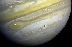 Le fond de l'image est occupé par Jupiter. Deux sphères sont devant la planète, une orangée (Io) vers la gauche et une grisâtre (Europe) vers le centre.