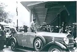 Photo ancienne représentant un convoi militaire, vu de dos, marchant dans une rue. On aperçoit des passants en arrière-plan.