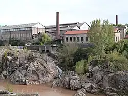 photo couleur montrant un chaos sur une rivière. Sur l'autre rive, plusieurs longs bâtiments industriels et deux hautes cheminées constituent une usine.