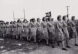 Un groupe de femmes en uniforme défile. Elles portent un petit étendard avec le symbole du corps des ingénieurs ; un château surmonté de la lettre D.