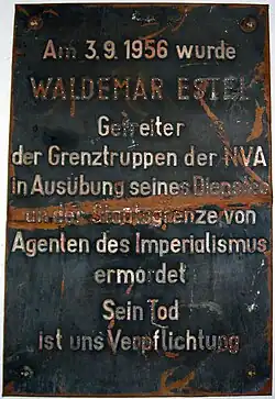Plaque de métal endommagée indiquant «&nbsp;Am 3.9.1956 wurde WALDEMAR ESTEL Getreiter der Grenztruppen der NVA in Ausübung seine Dienstes an der Staatsgrenze von Agenten des Imperialismus ermordet. Sein Tod ist uns Verpflichtung.&nbsp;»