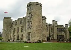 Photographie moderne d'un château en pierre comportant deux grandes tours rondes et un fronton sur lequel le drapeau de l'Angleterre flotte au vent.