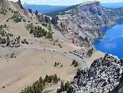 Vue plongeante depuis des rochers sur une route et un parking dominant le lac cerné par des reliefs déchiquetés.