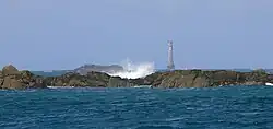 Au premier plan, rochers sur lesquels se fracasse une vague. Le phare est en arrière-plan, dans une légère brume.