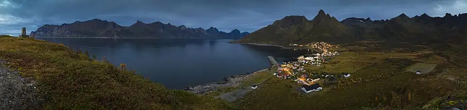 Posées sur une terre sombre, sous un ciel gris, au bord d'une eau noire, les maisons d'un village apparaissent comme des petites étoiles.