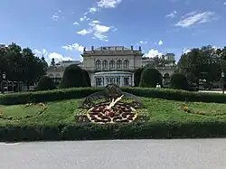 Des fleurs dessinent une horloge que des aiguilles parcourent. Derrière un grand bâtiment style demeure XIXème.
