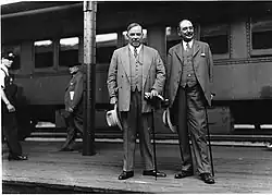 Photographie noir et blanc de deux hommes debout sur un quai avec un train derrière.