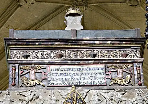 Photo en contre-plongée d'un coffre oblong avec des décorations florales dorées. Au centre, une plaque tenue par deux anges porte une inscription latine en majuscules. Une couronne en or est posée au sommet du coffre.