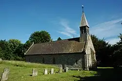 Photo d'un bâtiment allongé aux murs de pierres grises avec un petit clocher surmonté d'une girouette. Une rangée de pierre tombales apparaît dans l'herbe au premier plan.