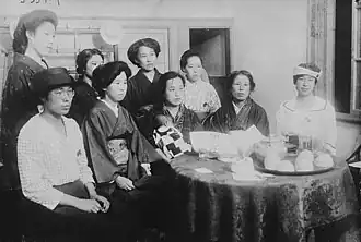 Photo de neuf femmes assises autour d'une petite table ronde. Certaines d'entre elles portent des vêtements traditionnels japonais, d'autres, des vêtements à la mode occidentale.