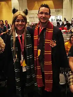 Un homme et une femme, tous deux à lunettes, posant l'un à côté de l'autre, portant notamment une écharpe rouge et jaune pour l'homme brun à droite et une écharpe verte et grise pour la femme blonde à gauche.