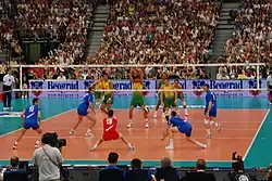 vue de derrière d'un mathc de volley-ball: au premier plan, la Serbie, avec un libéro en rouge qui s'apprête à réaliser une réception