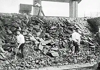 Photo en noir et blanc montrant des hommes travaillant sur une pente de pierre, au dessus un homme pousse un wagonnet.