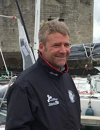 En buste, de trois quarts, dans un port, au pied d'une tour moyenâgeuse.