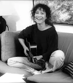 Photographie en nuances de gris de Yehudit Ravitz jouant de la guitare acoustique sur un canapé