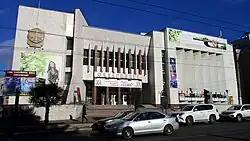 Bâtiment brutaliste avec une façade blanche ornée de panneaux publicitaires et des éléments géométriques marquéss. Devant, des voitures sont stationnées le long de la rue sous un ciel dégagé.
