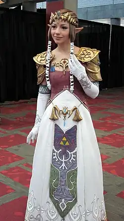 Femme blonde sur un fond noir, vêtue d'une robe blanche et violette et d'une ceinture, c'est un Cosplay de la Princesse Zelda.