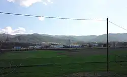 Au fond de la photo, une petite zone artisanale, avec quelques hangars métalliques ; au premier plan, grand champ verdoyant et poteaux téléphoniques. En arrière-plan, des collines.