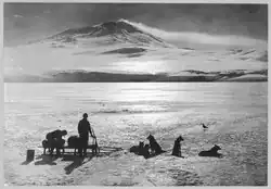 Photo en noir et blanc de deux hommes et cinq chiens attachés à un traîneau à l'arrêt sur la banquise devant un volcan.