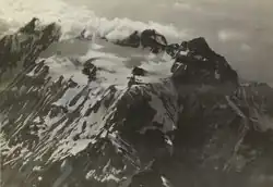 Photo ancienne d'une montagne sombre dans les nuages avec un glacier blanc posé aux pieds des pics.