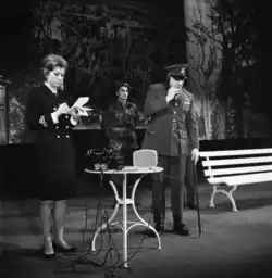 Une image de la scène de la production donnée au Stadttheater Bern où l'on voit trois personnes debout, l'une lisant un document et un autre débout au téléphone. Les trois portent des habits militaires.