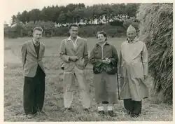 Groupe de quatre personnes dans un champs, avec une botte de foin sur la droite et une forêt en arrière-plan. Jacoba Hol est au centre de la photo.