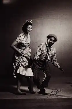 Photographie en noir et blanc, une femme est debout à gauche et à droite un homme est légèrement accroupi. La femme est vêtue d'une robe et l'homme d'un pantalon et d'une chemise.