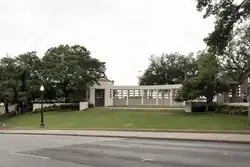 Une construction blanche sur un monticule gazonneux, entourée d'arbres. Au premier plan une route
