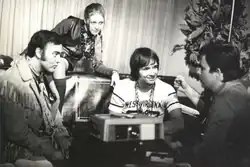 Photographie en noir et blanc de trois hommes assis. L'homme du milieu est assis sur un canapé, une femme est assise sur le dessus de ce meuble et est penchée vers lui.